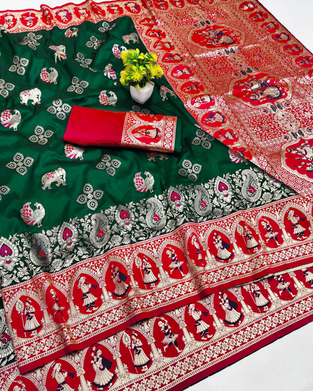 A green and red Banarasi silk saree with intricate gold zari embroidery, featuring elephant motifs on the body and detailed borders, styled with a red and gold matching blouse piece and a small vase of yellow flowers.
